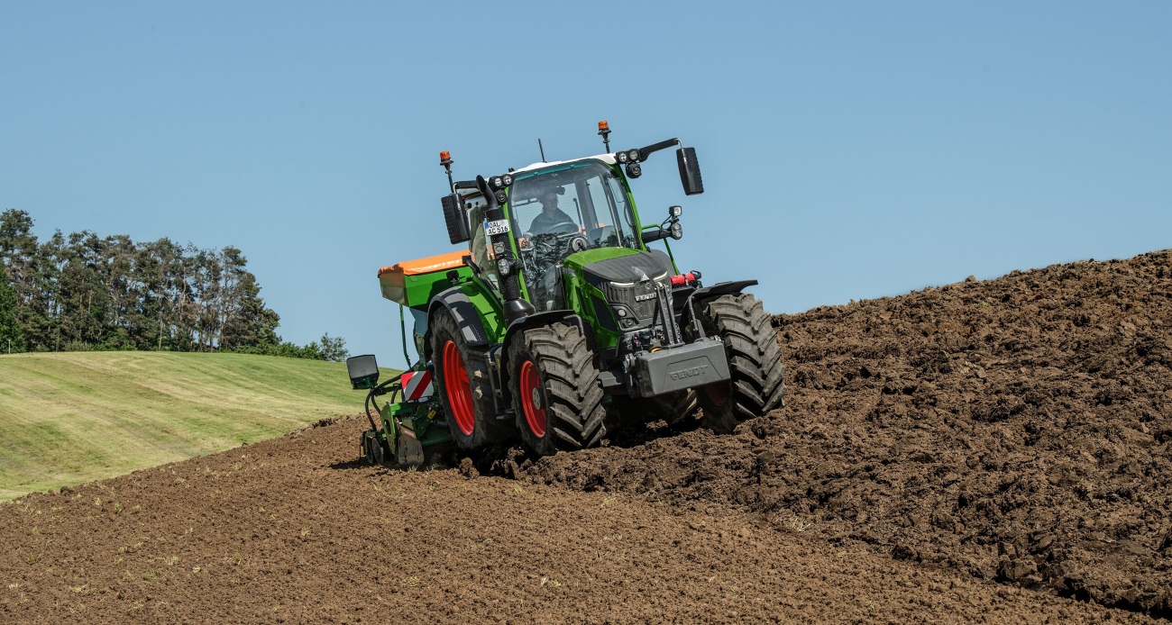 Ένα πράσινο Fendt 500 Vario διασχίζει ένα χωράφι με συνδυασμό σπαρτικής μηχανής από την Amazone σε πράσινο-πορτοκαλί χρώμα και σπορά.