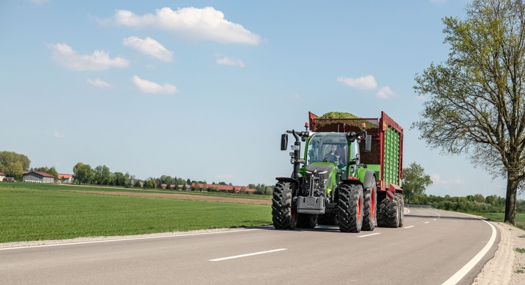 Ένα πράσινο Fendt 500 Vario οδηγεί με ένα γεμάτο βαγόνι μεταφοράς ζωοτροφών σε έναν δρόμο προς την κάμερα.