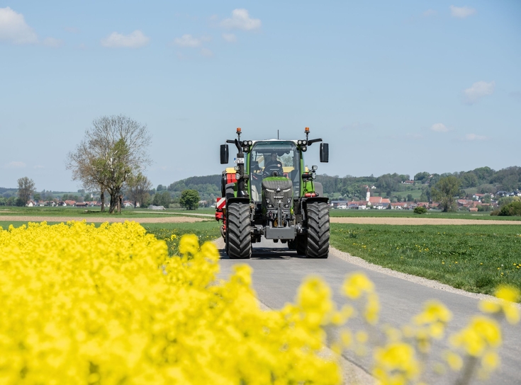 Ένα Fendt 500 Vario κινείται δίπλα σε ένα χωράφι με ελαιοκράμβη στο δρόμο και μεταφέρει ένα εργαλείο στο πίσω μέρος.
