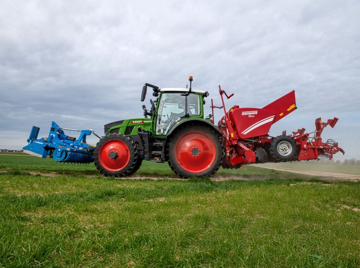 Ένα πράσινο Fendt 500 Vario διασχίζει έναν χωματόδρομο με μια κόκκινη σπαρτική μηχανή πατάτας στο πίσω μέρος και μια μπλε δισκοσβάρνα μπροστά.
