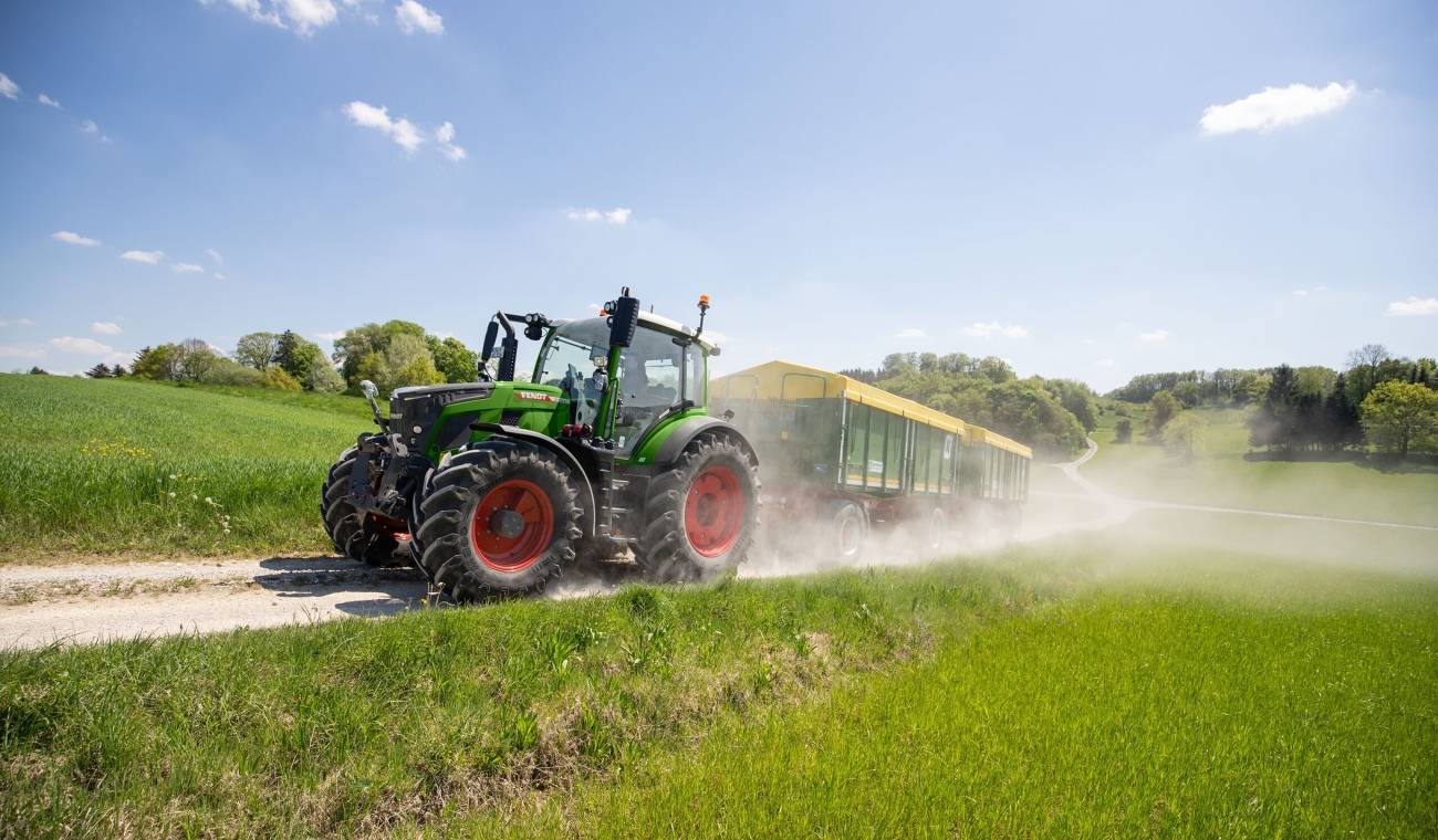Ένα πράσινο Fendt 500 Vario ρυμουλκεί δύο πράσινα τρέιλερ με κίτρινο κάλυμμα αλουμινίου πάνω σε χωματόδρομο και στροβιλιζόμενη σκόνη.