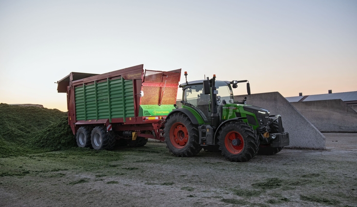 Ένα πράσινο Fendt 500 Vario ξεφορτώνει ένα πρασινοκόκκινο ρυμουλκούμενο Strautmann το σούρουπο σε ένα επίπεδο σιλό.