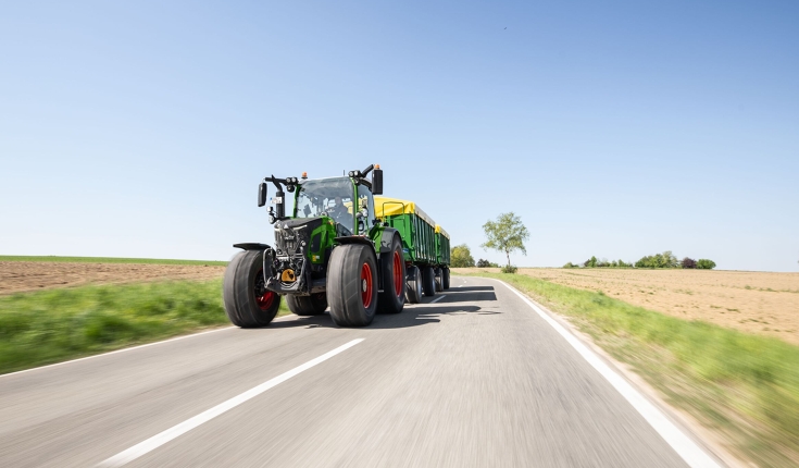 Ένα πράσινο Fendt 500 Vario κινείται σε έναν δρόμο με δύο πράσινα ρυμουλκούμενα με κίτρινο κάλυμμα από αλουμινόχαρτο.